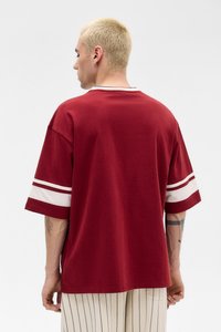 Maglietta rossa in cotone con dettagli a righe bianche sulle maniche, vestibilità rilassata, scollo tondo e orlo dritto. Presenta un design semplice sul retro.