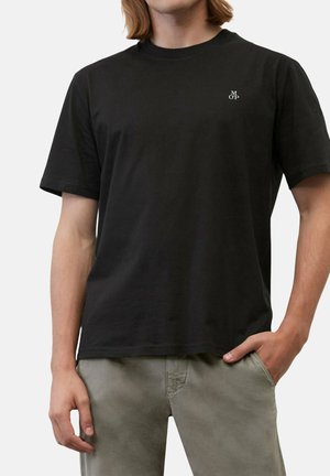 Jeune personne portant un t-shirt noir uni à manches courtes avec un petit logo blanc « M OP » sur la poitrine, associé à un pantalon gris clair.