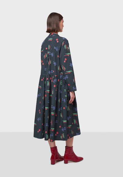 Vestido de manga larga oscuro con patrones geométricos coloridos en verde, rojo y azul. Falda acampanada, cintura ajustada, acompañado de botas rojas hasta el tobillo.