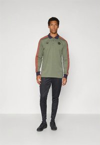 Olivengrønn langermet poloshirt med marineblå krage, røde striper på ermene, og svarte bukser med hvite sidestriper. Sko med gummisåle.