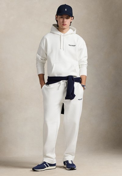 Hvit hettegenser med "POLO SPORT"-logo, hvite joggebukser med marineblå sjal bundet rundt livet, og marineblå caps. Marineblå og hvite sportssko.