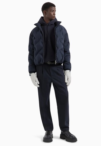 Giacca puffer blu navy con colletto alto, abbinata a una felpa scura, pantaloni sartoriali e guanti chiari, con scarpe lace-up nere.