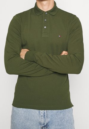 Osoba ubrana w oliwkowo zieloną koszulkę polo z długim rękawem i trzema guzikami, z założonymi rękami, w jasnoniebieskich dżinsach widocznych w talii.