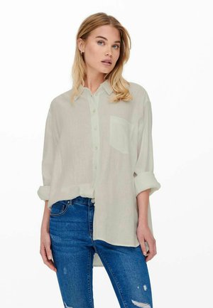 Light green button-up shirt made of a soft, sheer fabric with rolled sleeves and a chest pocket, worn over blue distressed jeans.
