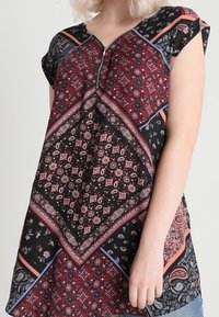 Tmavý top s výstřihem do V, krátkými rukávy a složitými paisley vzory v červené, černé a krémové barvě. Obsahuje centrální zipový detail.