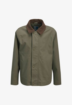 Smilende dreng iført rød kasket, grøn jakke, ternet skjorte, hvid T-shirt, løse blå jeans og beige sko, stående med den ene hånd i lommen.