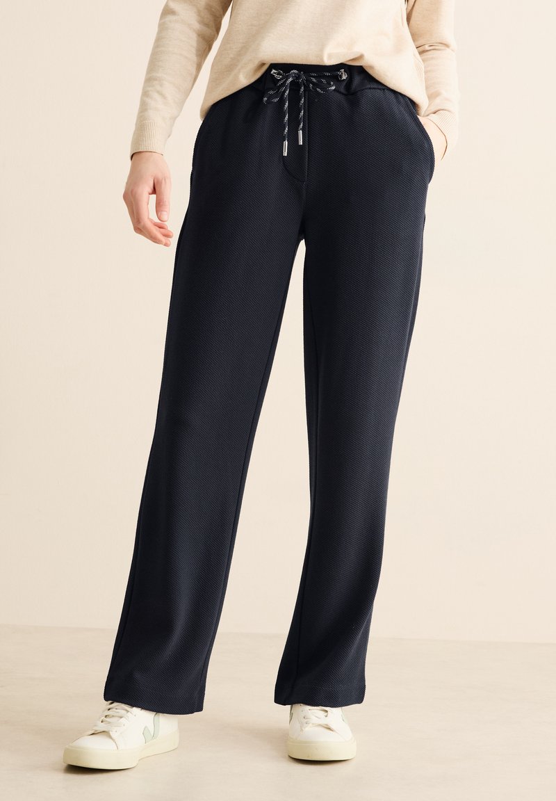 Pantalons amples de couleur marine en tissu texturé avec taille à cordon réglable, poches latérales, et associés à des baskets blanches.