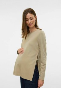 Maglietta premaman beige a maniche lunghe con un fit rilassato, legature laterali e tessuto morbido. Abbinata a jeans scuri, evidenziando il design della maglietta.