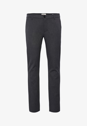 Mann trägt einen grauen Langarmpullover, dunkle eng geschnittene Hose und schwarze Lederloafer und steht vor einem einfarbigen Hintergrund.