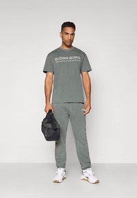 Vīrietis pelēkā Björn Borg T-kreklā un sporta biksēs, tur melnu sporta somu, stāvot pret viegliem fona, uz kājām baltas kedas.