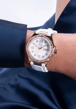 Silver and rose gold watch with a white textured dial, gemstone accents, and a white rubber strap, displaying Roman numerals and date function.