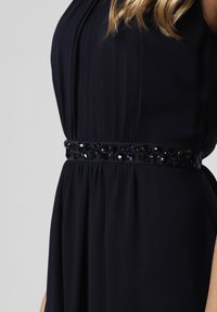 Kvinna klädd i en svart klänning med rynkad tyg och ett dekorativt svart pärlarmband i midjan, mot en ljusgrå bakgrund.