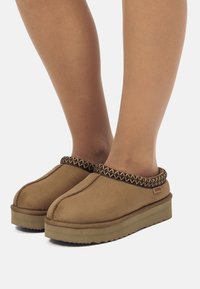 Zapatos marrones tipo slip-on con un exterior texturizado, que presentan un ribete trenzado con patrón y una suela gruesa y duradera.