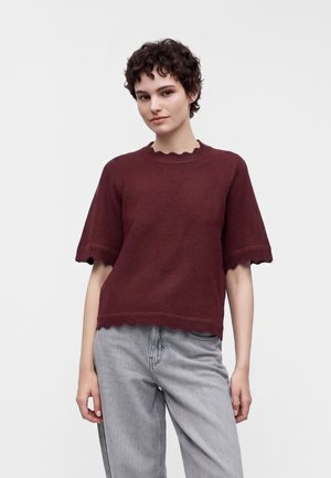Femme aux cheveux courts et bouclés portant un pull bordeaux à manches courtes avec des bords festonnés et un jean taille haute gris clair, debout devant un fond uni.