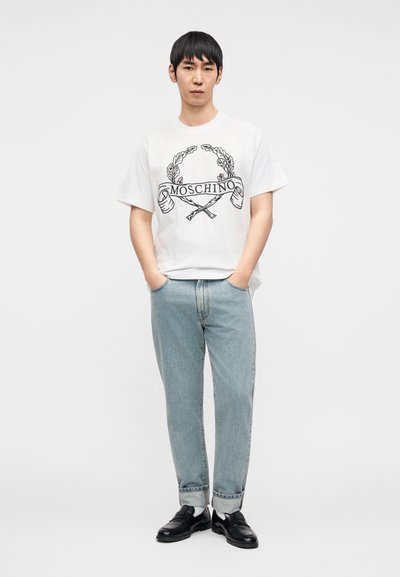 Άσπρη κοντομάνικη μπλούζα με μαύρο λογότυπο και γραφικό σχέδιο "Moschino", συνδυασμένη με ανοιχτό μπλε τζιν παντελόνι και μαύρα loafers.