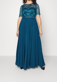 Femme portant une robe longue bleu sarcelle avec un corsage en dentelle florale, des manches courtes transparentes et une ceinture ornée de perles, debout devant un fond uni.