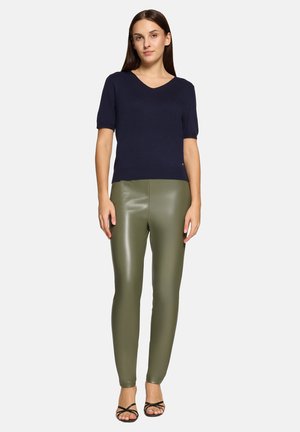 Frau steht, trägt ein marineblaues Kurzarmoberteil, olivgrüne Leggings aus Kunstleder und schwarze, offene Sandaletten mit Riemchen vor hellem Hintergrund.