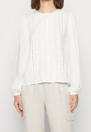 Kobieta ubrana w białą bluzkę z długim rękawem, ozdobioną pionowymi plisami i haftowanymi detalami, w zestawieniu z jasnobeżowymi spodniami z bocznymi kieszeniami.