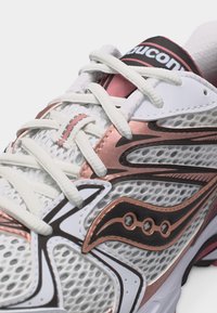 Zapatilla de correr de malla en blanco y oro rosa con cordones blancos y logo negro en el lateral y la lengüeta.