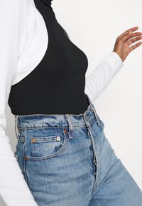 Schwarzer Einteiler kombiniert mit hochgeschnittenen blauen Jeans. Der Einteiler hat eine figurbetonte Silhouette, während die Jeans leichte Verzerrungen und Kupferakzente aufweisen.