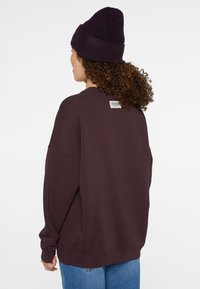 Dark purple oversized sweatshirt with ribbed cuffs and collar, featuring a sewn label at the back, paired with light blue denim jeans.