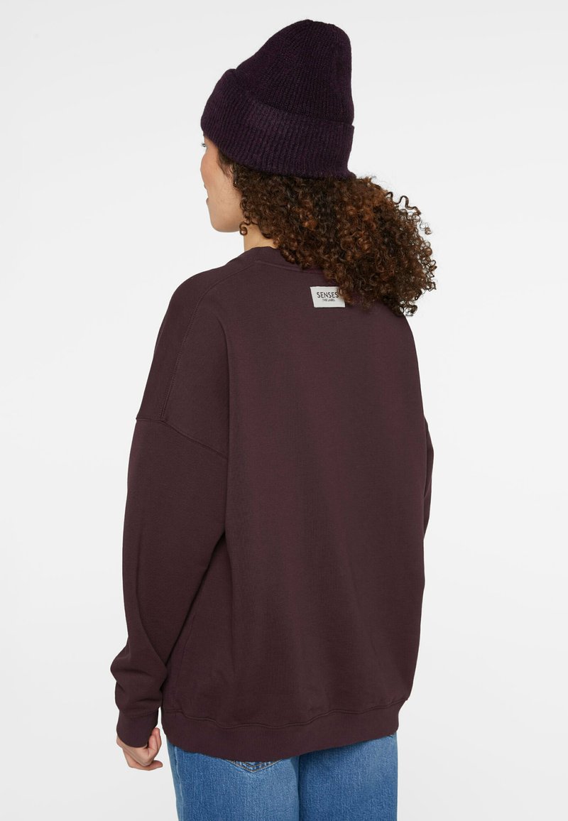 Dark purple oversized sweatshirt with ribbed cuffs and collar, featuring a sewn label at the back, paired with light blue denim jeans.