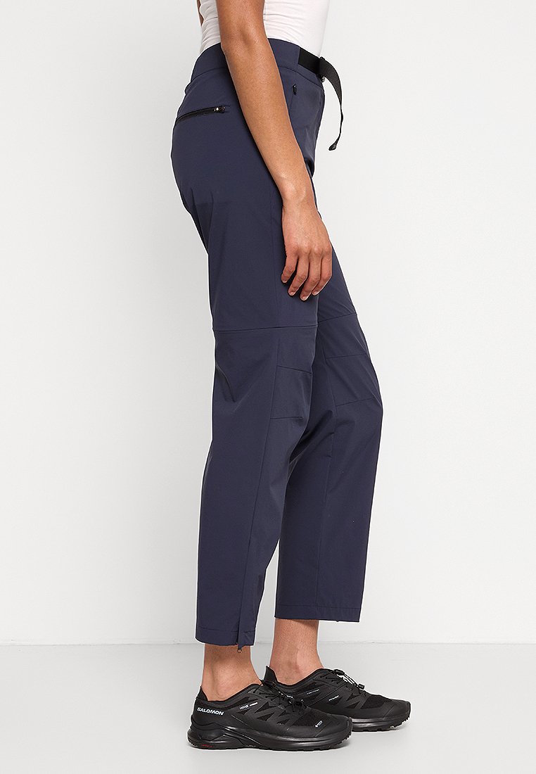 Person trägt marinefarbene, knöchellange Hose mit Reißverschlusstasche und schwarze Sportschuhe, steht vor einem schlichten hellen Hintergrund.