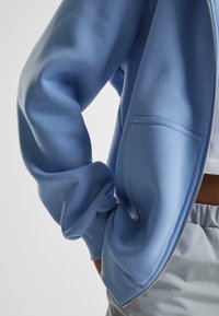 Felpa leggera blu ceruleo con zip, dotata di polsini e orlo a coste. Presenta una tasca frontale e una texture di tessuto liscia e morbida.