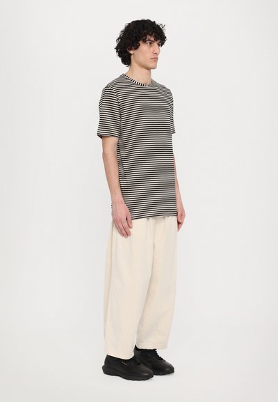 Un tricou raiat alb și negru cu mâneci scurte, combinat cu o pereche de pantaloni lărgi de culoare crem și pantofi negri. Fundal simplu.
