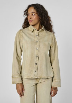 Frau mit langen lockigen Haaren, die eine beigen Knopfleisten-Jeansjacke und passende Hose trägt, schaut nach links vor einem schlichten Hintergrund.