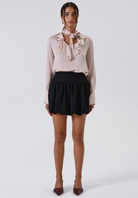 Donna che indossa una blusa rosa chiaro con rouches e cravatta al collo, una mini gonna nera a pieghe e scarpe con il tacco a punta color borgogna, in piedi contro uno sfondo bianco.