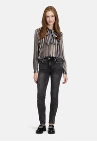 Vrouw met lang rood haar, gekleed in een gestreepte blouse met een strik, zwarte studs skinny jeans en glanzende zwarte loafers, staande op een witte achtergrond.