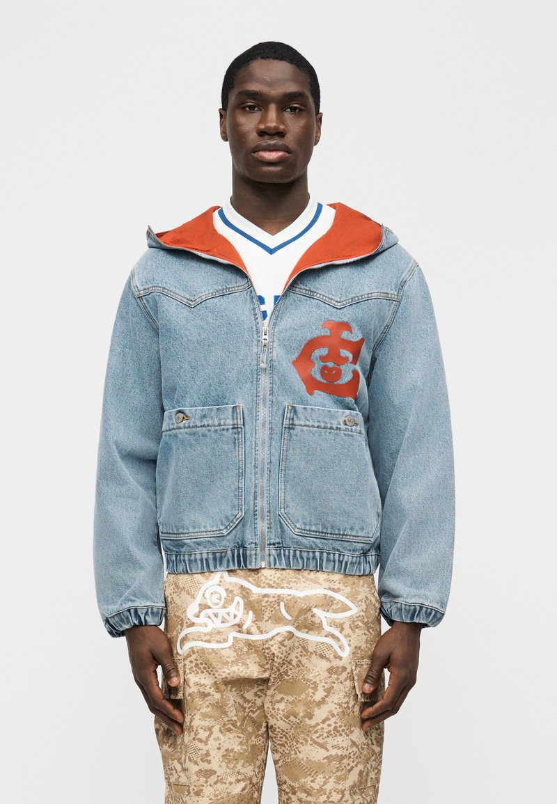 Jaunas vyras dėvi mėlynę džinsinę striukę su užtrauktuku, oranžine apdaila ir emblema, baltą marškinius ir smėlio spalvos raštuotas kelnes su baltu gyvūnų grafiniu piešiniu.