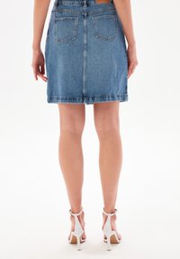 Kvinna stående, klädd i en blå denimkjol och vita högklackade sandaler, sedd bakifrån mot en vit bakgrund.