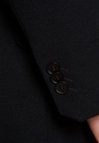 Rękaw ciemnej wełnianej marynarki z trzema czarnymi guzikami, każdy z czterema otworami, wykończony brązowym haftem. Gładka tekstura z dopasowanym krojem.