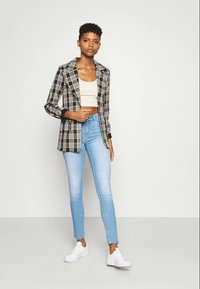 Μαύρο και άσπρο καρό σακάκι με μακριά μανίκια, φορεμένο πάνω από φ fitted κρεμ τοπ, συνδυασμένο με ανοιχτό μπλε skinny τζιν και λευκά αθλητικά παπούτσια.