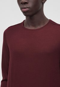 Jeune personne portant un t-shirt uni bordeaux à manches longues et col rond sur un fond gris neutre.