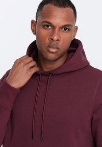 Felpa borgogna con cappuccio regolabile, dotata di cuciture piatte e una consistenza morbida. Design semplice, maniche lunghe e senza loghi visibili.