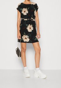 Czarna sukienka w kwiatowy wzór z krótkimi rękawami, zaciśnięta w talii i o luźnym kroju; w połączeniu z białymi, grubymi sneakersami i małą torebką.