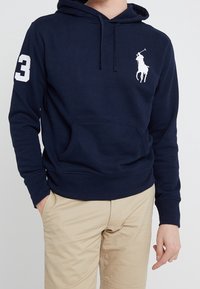 Osoba na sobě má tmavě modrou mikinu s kapucí, na hrudi s bílým logem póla a číslem 3 na rukávu, v kombinaci s béžovými kalhotami.