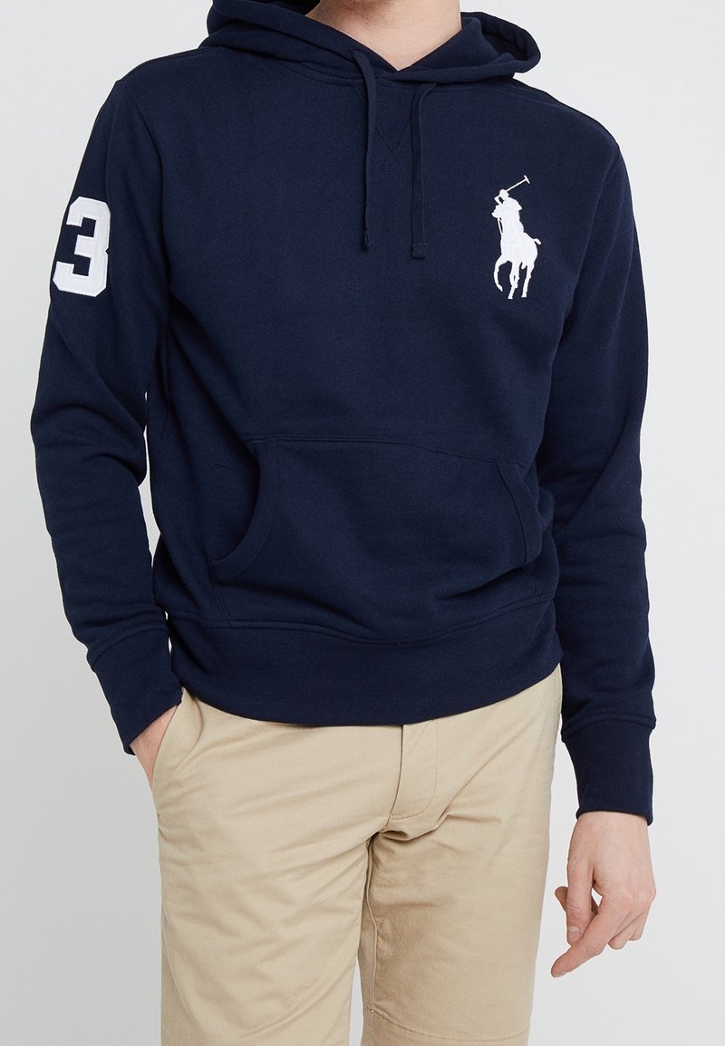 Osoba na sobě má tmavě modrou mikinu s kapucí, na hrudi s bílým logem póla a číslem 3 na rukávu, v kombinaci s béžovými kalhotami.