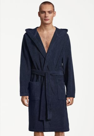 Mężczyzna w granatowym szlafroku z kapturem, przednimi kieszeniami i zawiązanym paskiem w pasie, stojący na tle jednolitego białego tła.
