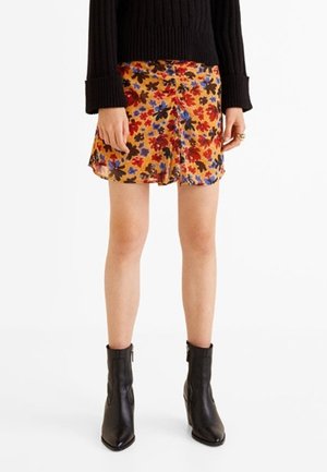 Kobieta ubrana w żółtą spódnicę w kwiaty, czarny prążkowany sweter i czarne botki, stojąca na tle białego tła.