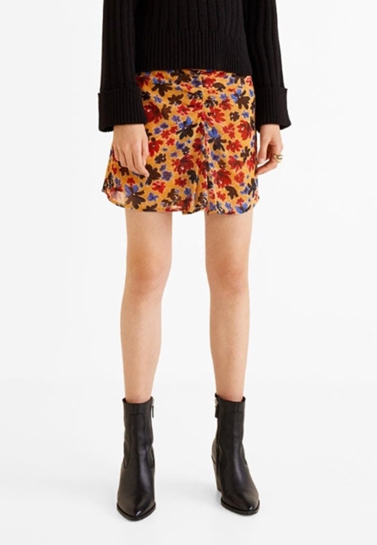 Femme portant une jupe jaune fleurie, un pull noir côtelé et des bottines noires, debout sur un fond blanc.