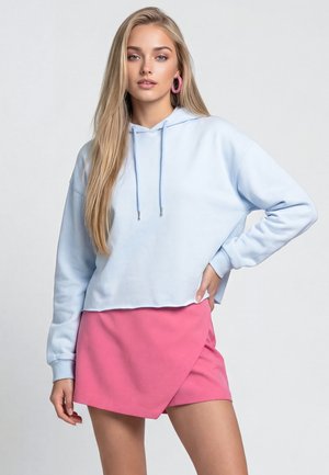 Jonge vrouw met lang blond haar, die een lichtblauwe hoodie, een roze rok en grote roze oorringen draagt, staand tegen een effen achtergrond.
