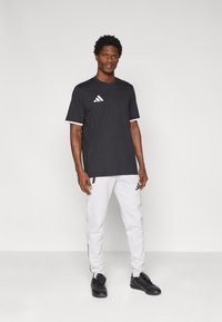 Μαύρο T-shirt με κοντά μανίκια και λευκές λεπτομέρειες, με λογότυπο στο αριστερό στήθος. Συνδυασμένο με λευκό παντελόνι και μαύρα αθλητικά παπούτσια.