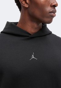 Gros plan d'une personne portant un sweat à capuche noir avec un petit logo argenté Jumpman centré sur la poitrine.