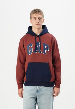 Giovane uomo che indossa un hoodie bordeaux e blu navy con il logo "GAP" e jeans azzurri, in piedi contro uno sfondo bianco semplice.