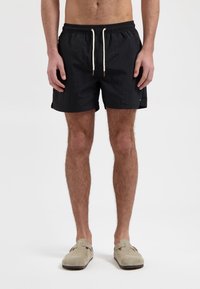 Schwarze Badehosen mit elastischem Bund und Kordelzug. Gewebter Stoff, bis zur Mitte des Oberschenkels, mit Seitentaschen. Kombiniert mit hellgrauen Slippers.