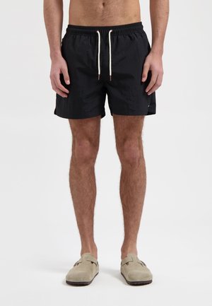 Schwarze Badehosen mit elastischem Bund und Kordelzug. Gewebter Stoff, bis zur Mitte des Oberschenkels, mit Seitentaschen. Kombiniert mit hellgrauen Slippers.
